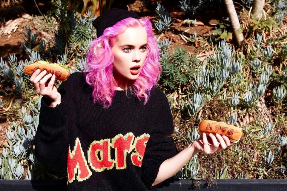 Young woman with pink hair poses outdoors holding two hotdogs with the word LOVE carved in each bun, wearing a black sweater and beanie.