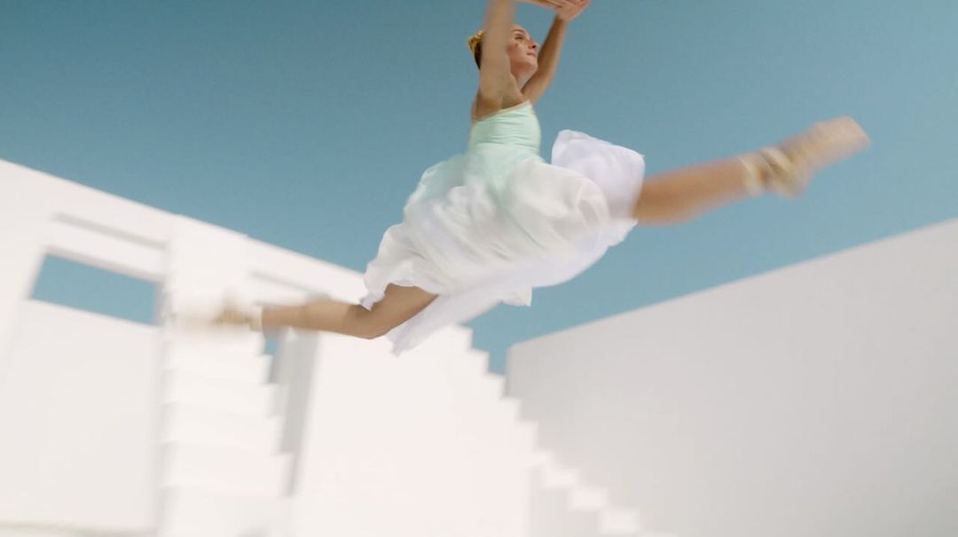 A young female ballet dancer is leaping in the air with legs split against a white staircase and light blue background, performing ballet.