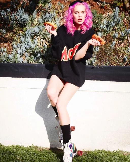 Young woman with pink hair balances on a skateboard outside, holding a hot dog in each hand, wearing a black AC/DC sweater.
