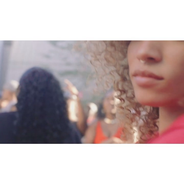 Close-up of an adult female with curly blonde hair and a nose ring, with a blurred crowd in the background at an outdoor event.