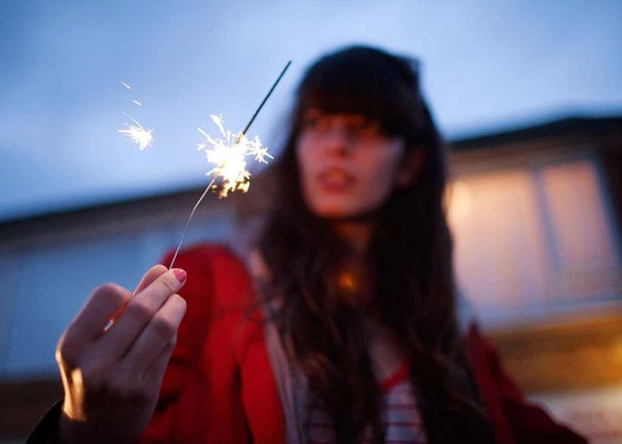 Close-up of a woman holding a sparkler at dusk, with bokeh lights and a blurred architectural backdrop. Celebratory and festive atmosphere.