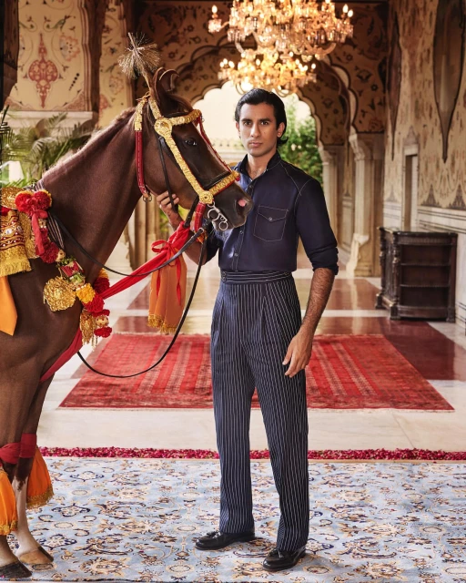 Man in vintage pinstripe pants poses with a decorated brown horse inside an ornate, old-world hall with patterned rugs and chandelier.