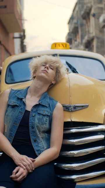 Woman rests on the hood of a yellow taxi with buildings visible in the background, creating an urban setting.
