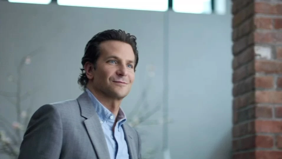 Portrait of adult male in a gray suit and light blue shirt, smiling indoors with light gray wall and brick column in the background.