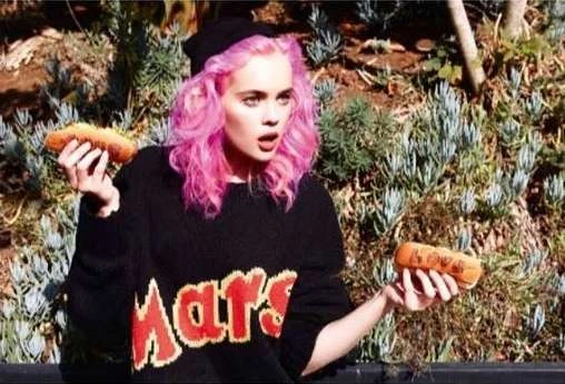 A young woman with pink hair and a black sweater holds two hot dogs outdoors. She appears surprised in a sunny, natural environment.