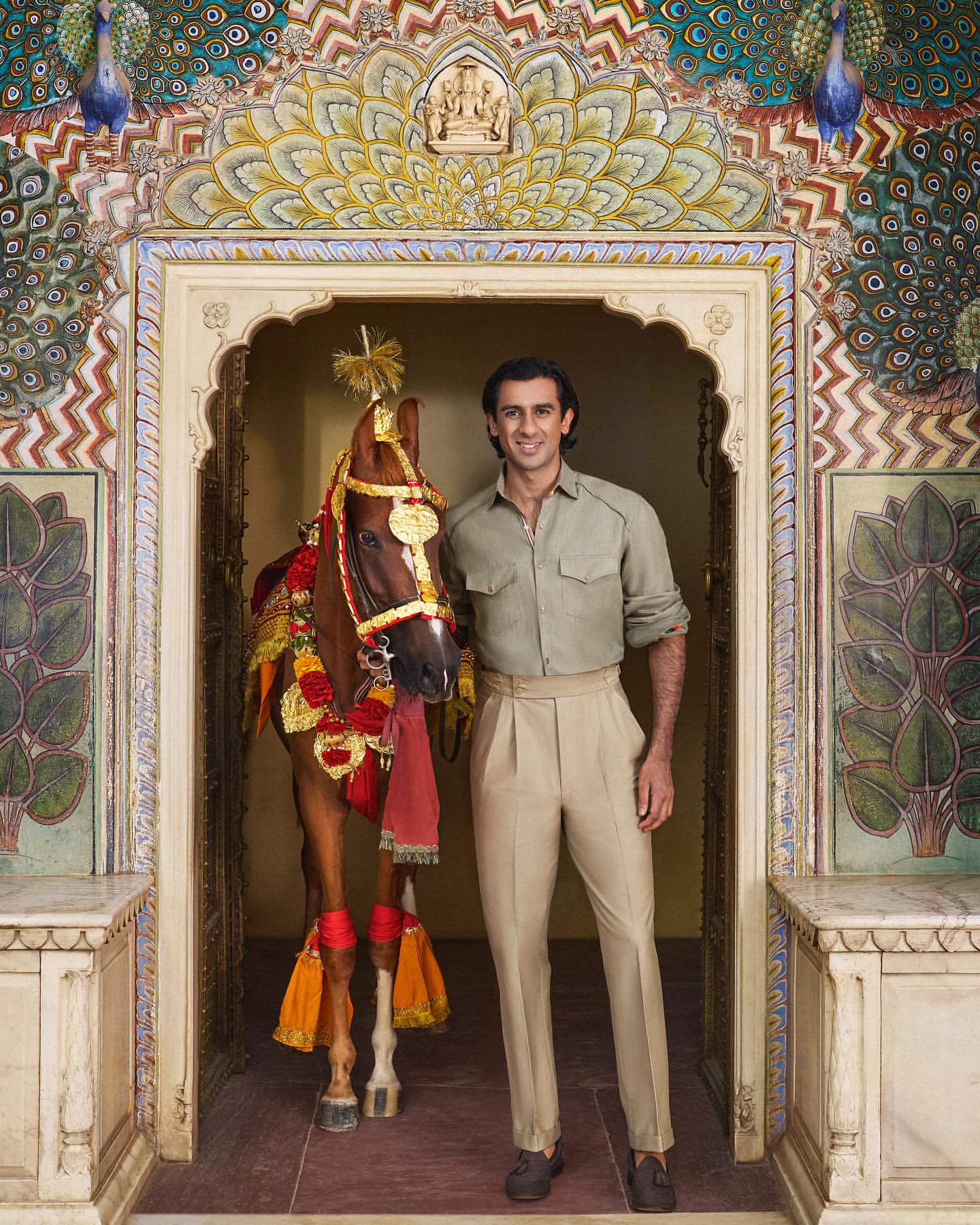 A man in casual attire stands next to a horse adorned with decorative tassels in front of an ornate palace archway with peacock designs.