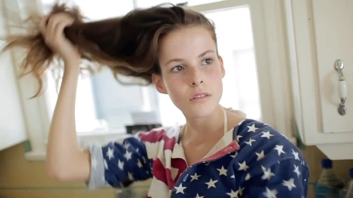 Young woman with long brown hair posing indoors, wearing a blue and white star patterned shirt, looking away from the camera.