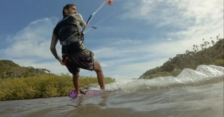 A man kite surfing on the ocean under a bright, cloudy sky, with a green, hilly coastline visible in the background and white waves.