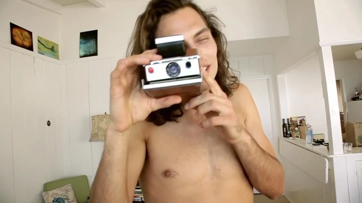Shirtless man with wavy dark hair holding a vintage Polaroid camera indoors, focusing on taking a photo with a neutral expression.