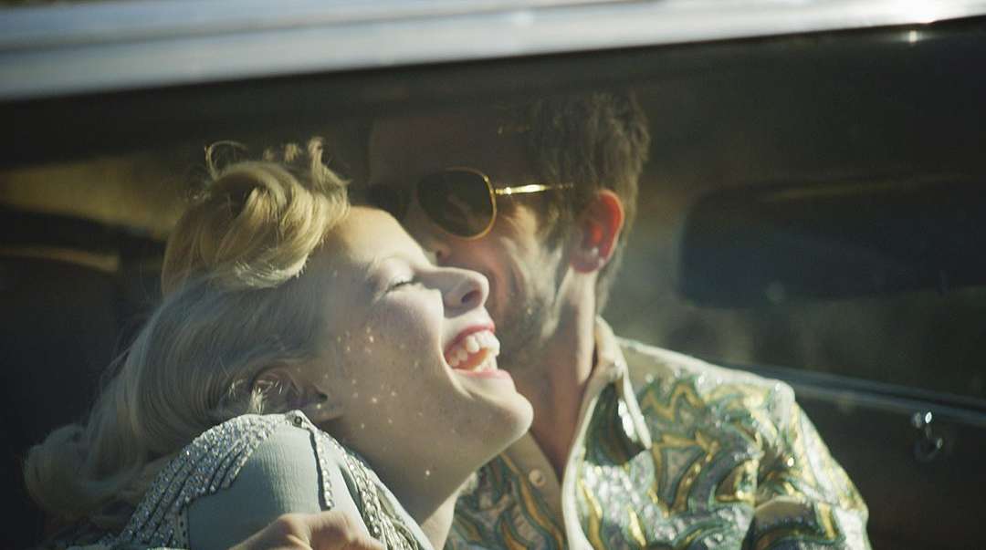 Close-up of a laughing woman with blonde hair and a man wearing sunglasses, captured inside a car with natural lighting.