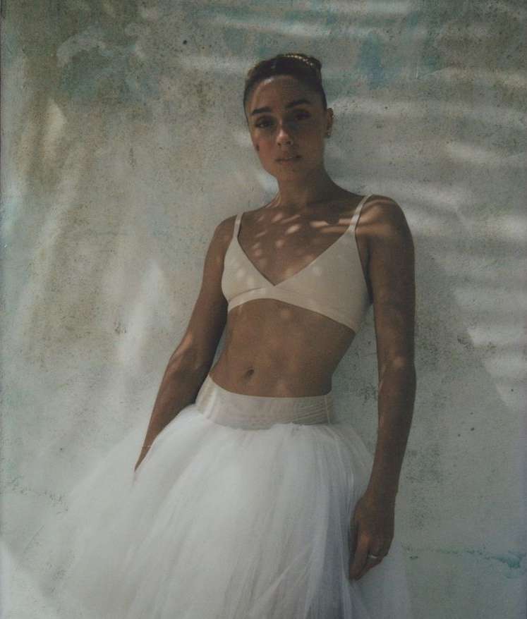 Portrait of a woman in a bra top and tutu skirt, posing against a textured wall in dappled sunlight.