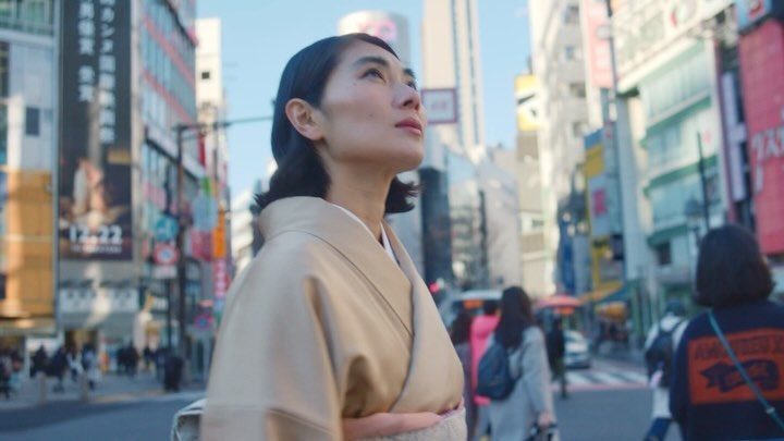 Woman in tan jacket stands in a sunny urban setting with modern buildings, pedestrians, and vibrant colors, showcasing street fashion.