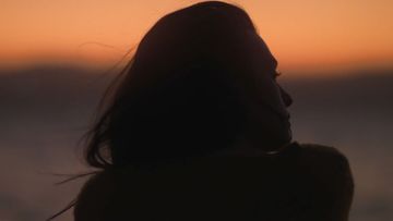 Silhouette of a woman gazing out at an orange sunset over the water, creating a serene and contemplative atmosphere.