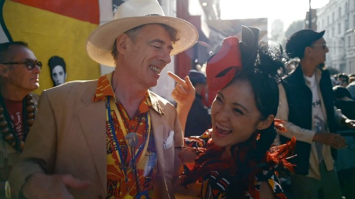 Two smiling people, an adult male in a hat and tan jacket, and an Asian adult female with a feathered headpiece, outdoors in daylight.