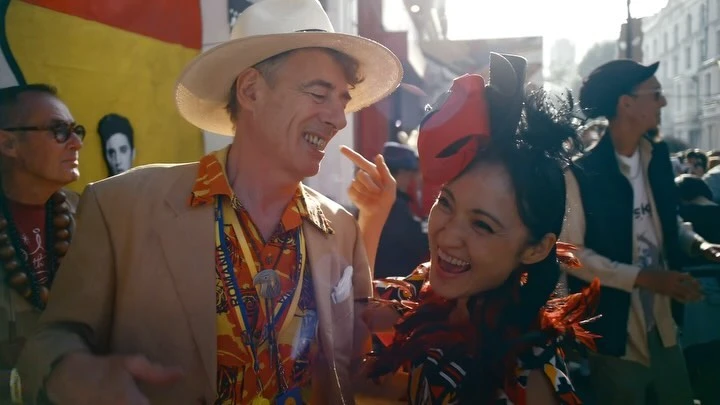 Two smiling people, an adult male in a hat and tan jacket, and an Asian adult female with a feathered headpiece, outdoors in daylight.