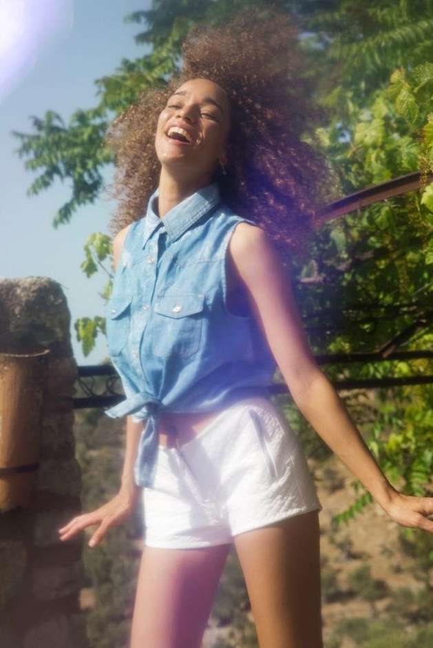 A young adult woman with curly hair smiles while wearing a denim top and white shorts, set against a sunny outdoor background with greenery.