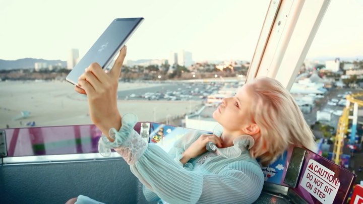 Young woman in a Ferris wheel cabin uses a tablet device with a cityscape, amusement park, beach, and ocean visible in the background.