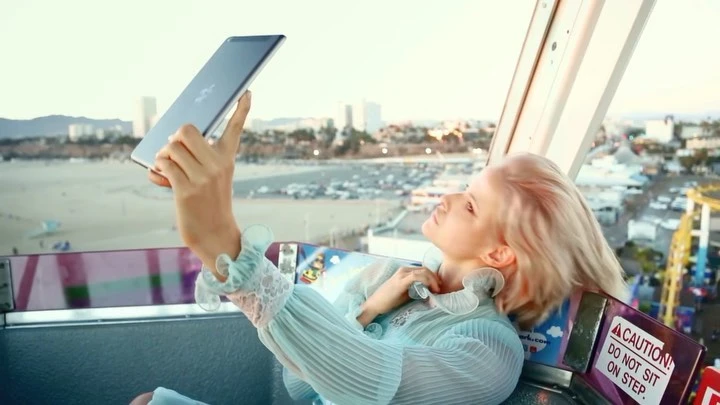 Young woman in a Ferris wheel cabin uses a tablet device with a cityscape, amusement park, beach, and ocean visible in the background.