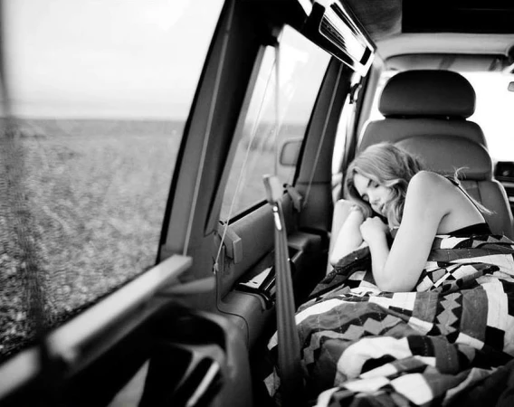 Monochrome interior photograph of a woman sleeping wrapped in a patterned blanket inside the back of a vehicle.