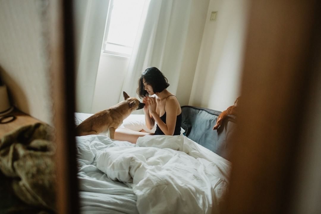 Woman in black tank top sitting on bed with a dog, natural light illuminates an indoor scene with white bedding and curtains.