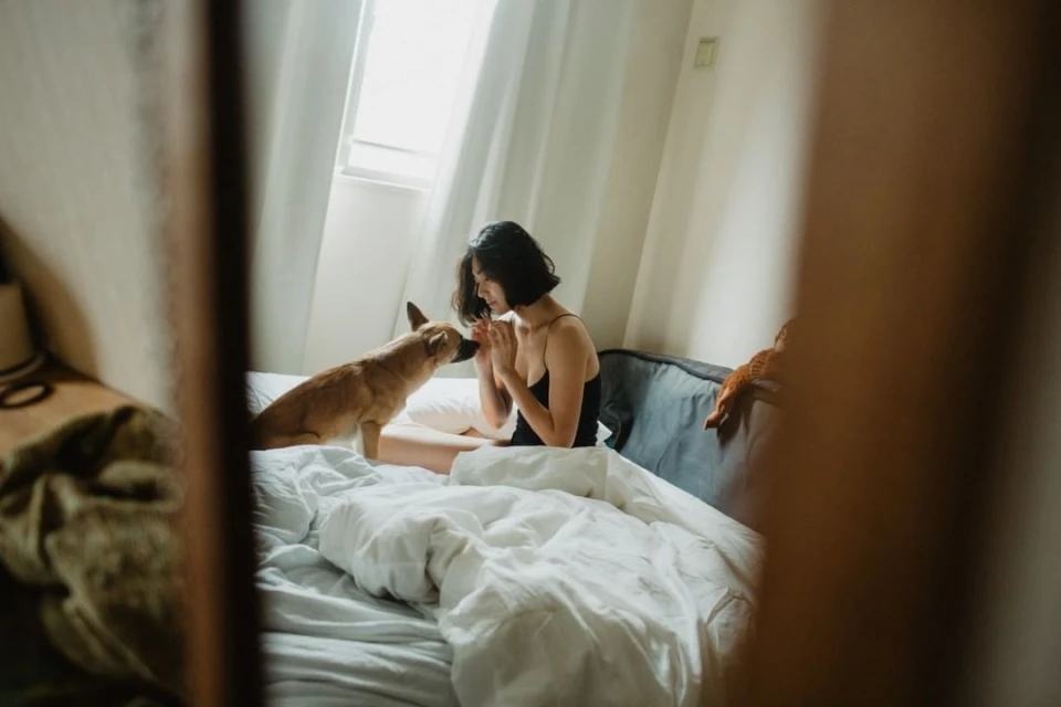 Woman in black tank top sitting on bed with a dog, natural light illuminates an indoor scene with white bedding and curtains.