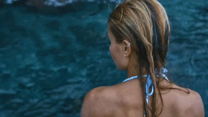 Rear view of a woman with wet blonde hair and small earrings wearing a light blue patterned halter top against dark water.