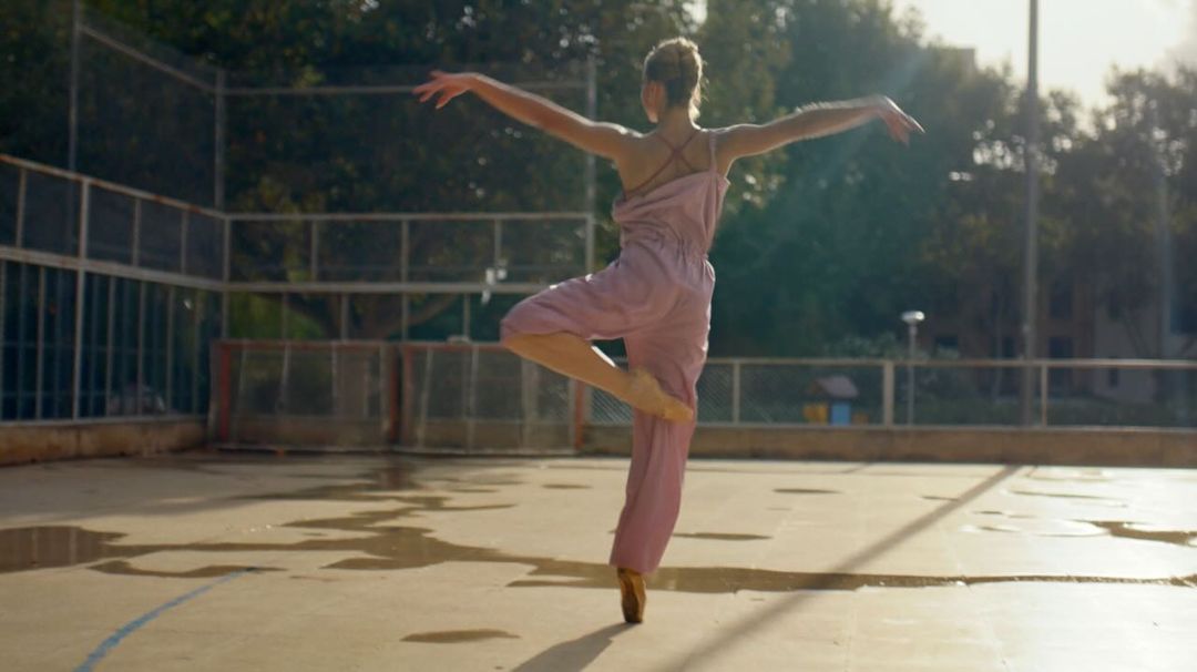 Solo ballet dancer in a light pink jumpsuit performs en pointe on a wet, outdoor court surrounded by trees and a chain link fence.