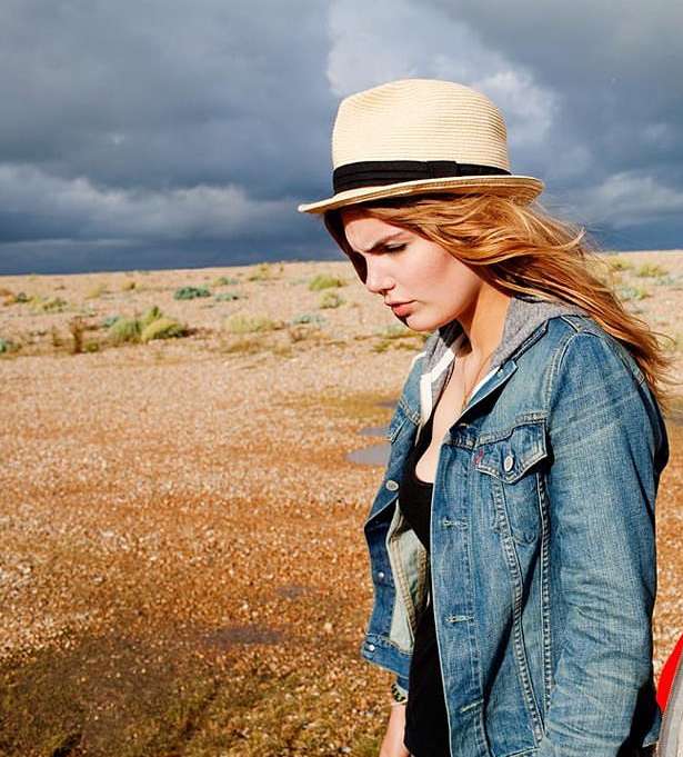 A young woman with strawberry-blonde hair stands on a beach with a cloudy sky. She wears a denim jacket and a straw hat.