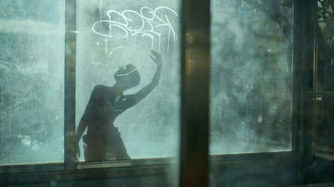 Silhouette of a person with an arm raised, seen through a dirty, graffitied window with outdoor greenery visible in the background.