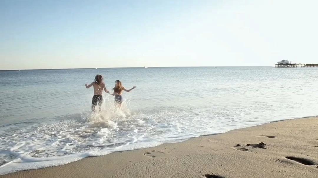Couple holding hands and running together in surf at the beach on a sunny day with a pier visible in the distance over the ocean.