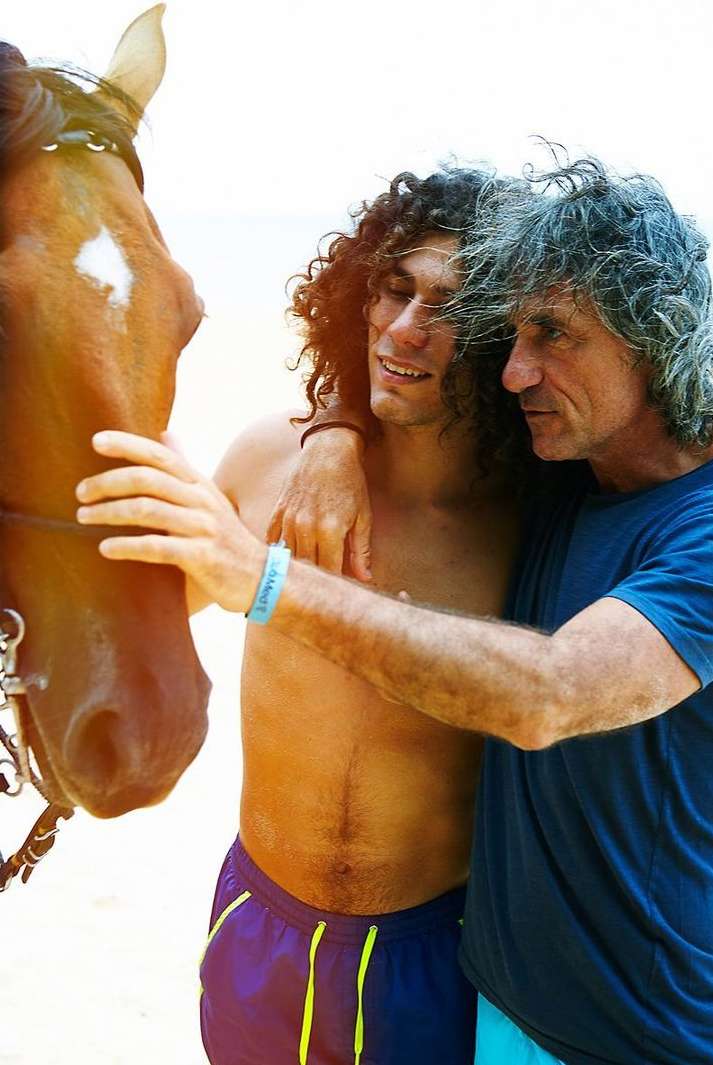 Two men bonding with a horse on a beach. The men, one older and one younger, stand close to the horse, with their arms around each other and the horse.