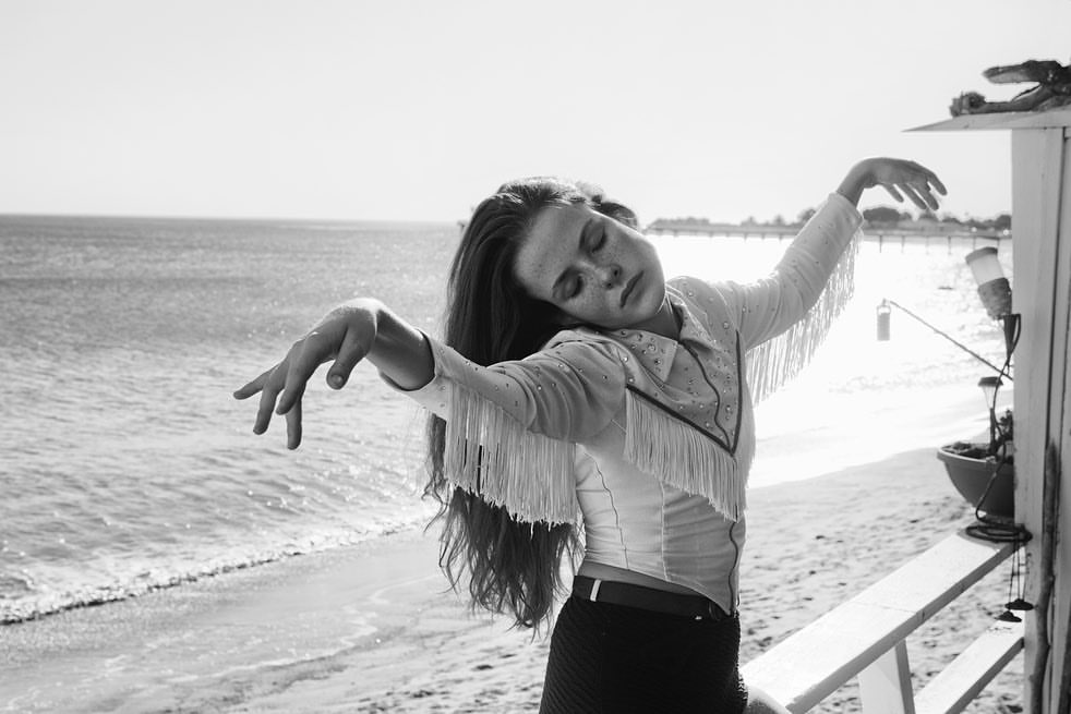 Young woman with long hair and freckles stands on a deck overlooking the ocean, wearing a fringed western-style shirt and dark skirt.