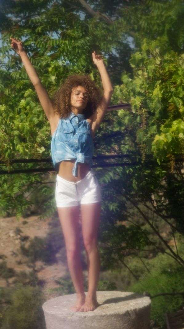 Young woman with curly hair and a tie-dye top stands barefoot on a concrete plinth outdoors, arms raised, surrounded by green foliage.