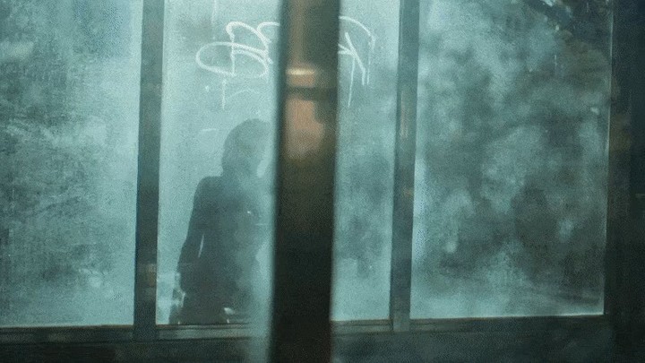 Silhouetted figure visible through a graffiti-covered glass pane. A dark figure appears indistinct, with a blue-green cast from the window glass.