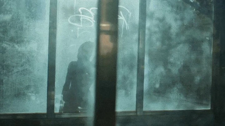 Silhouetted figure visible through a graffiti-covered glass pane. A dark figure appears indistinct, with a blue-green cast from the window glass.