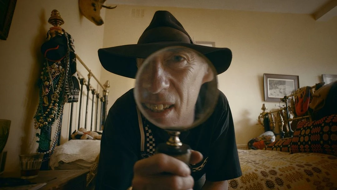Close-up of a man in a hat holding a magnifying glass in an interior setting, with visible objects in the background.