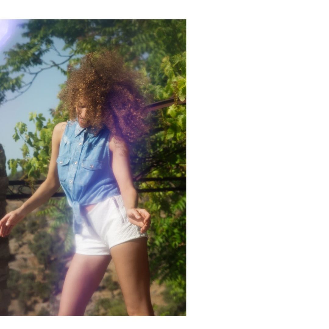 Stylish woman with curly hair dancing in sunlight wearing a denim shirt and white shorts with a green leafy tree background.
