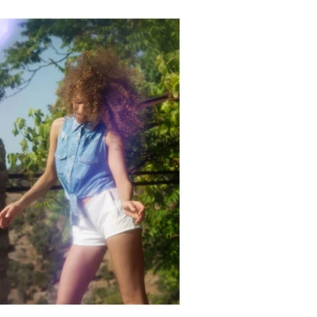 Stylish woman with curly hair dancing in sunlight wearing a denim shirt and white shorts with a green leafy tree background.