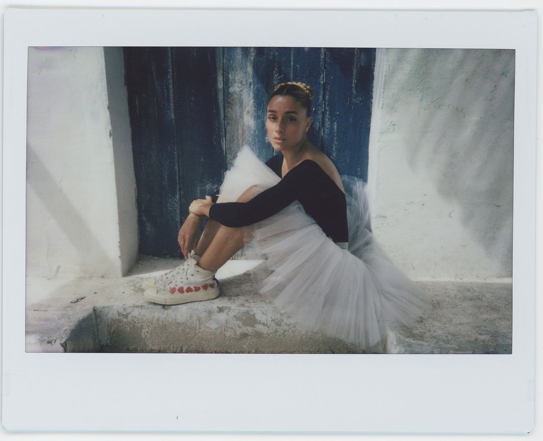 Young woman in ballerina style sits in front of weathered blue door. She's wearing a tutu and sneakers with red hearts.