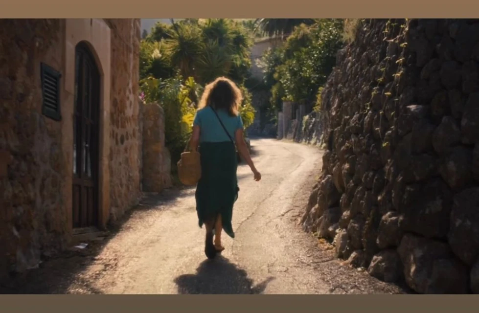 Back view of woman walking along a stone path lined with buildings and lush greenery in a European village on a sunny day.