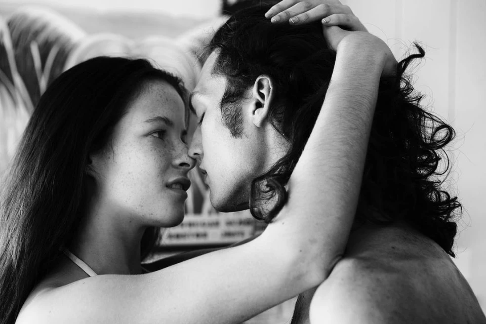 Black and white close-up portrait of a couple embracing with faces close, the woman has freckles, and the man has long curly hair.