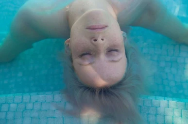 Close-up portrait of a woman with freckles floating on her back in a bright blue swimming pool with her eyes closed.