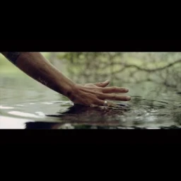Close-up of a person's hand touching the surface of calm water, creating ripples, captured in a cinematic widescreen composition.
