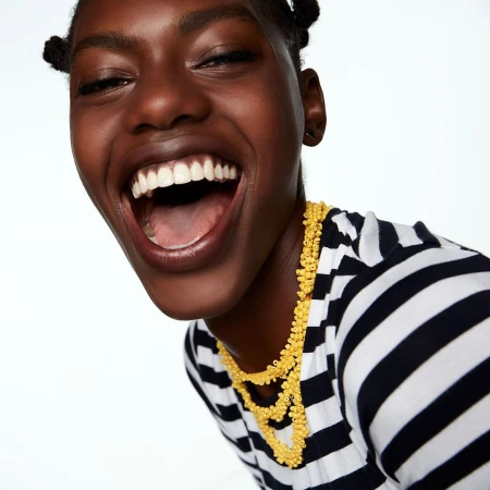 Close-up portrait of a laughing Black woman wearing a black and white striped top and a yellow beaded necklace against a white background.