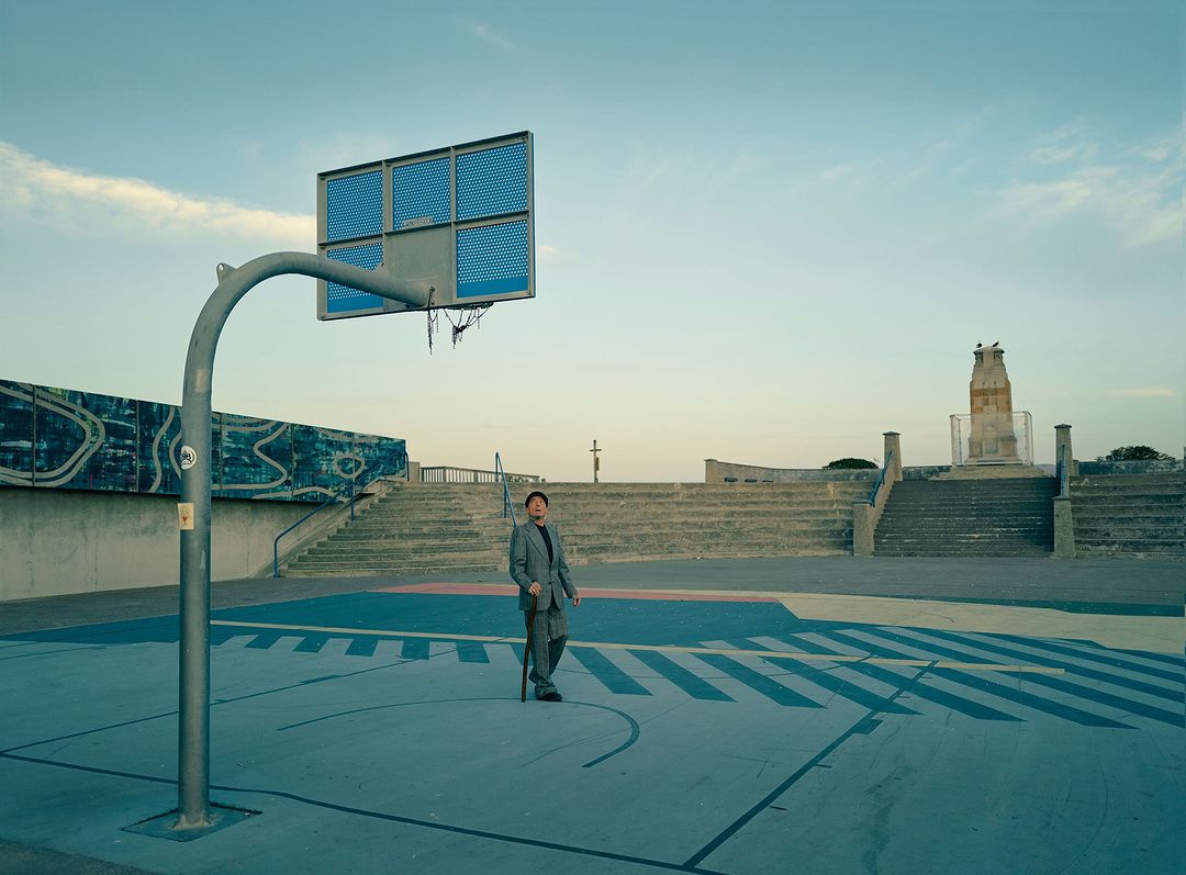 An adult man in a suit stands on an outdoor basketball court with a walking stick. The setting is bathed in soft, even light.