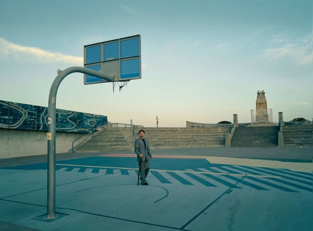 An adult man in a suit stands on an outdoor basketball court with a walking stick. The setting is bathed in soft, even light.