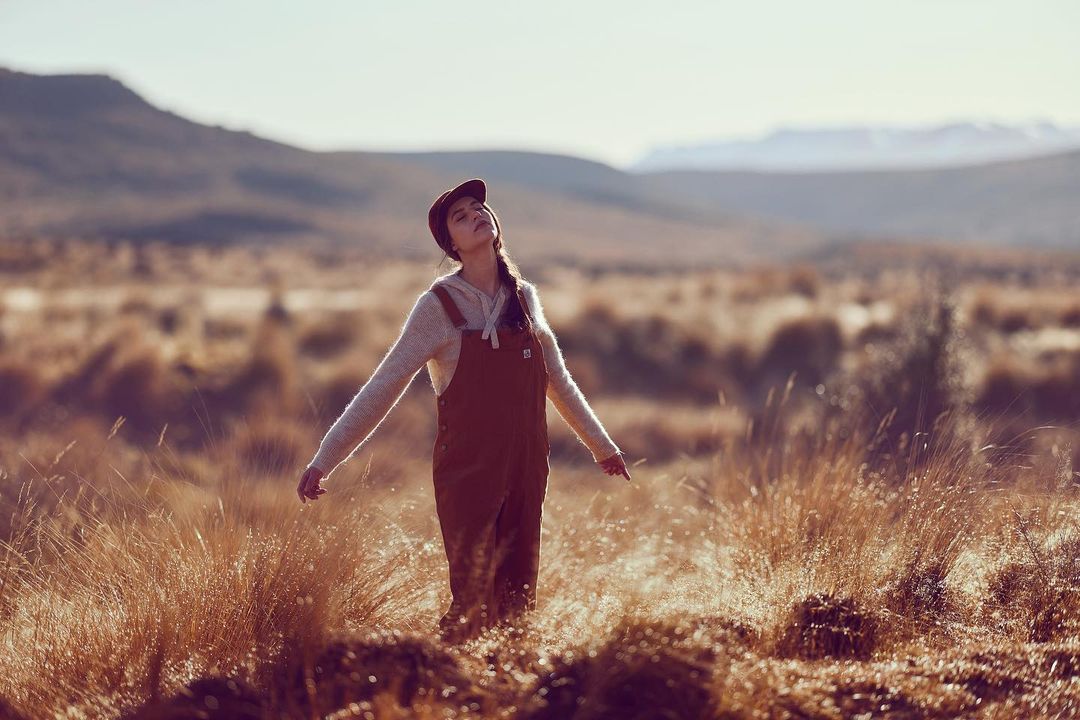 Woman in brown overalls and cap standing in a grassy field with arms outstretched under a soft hazy sky