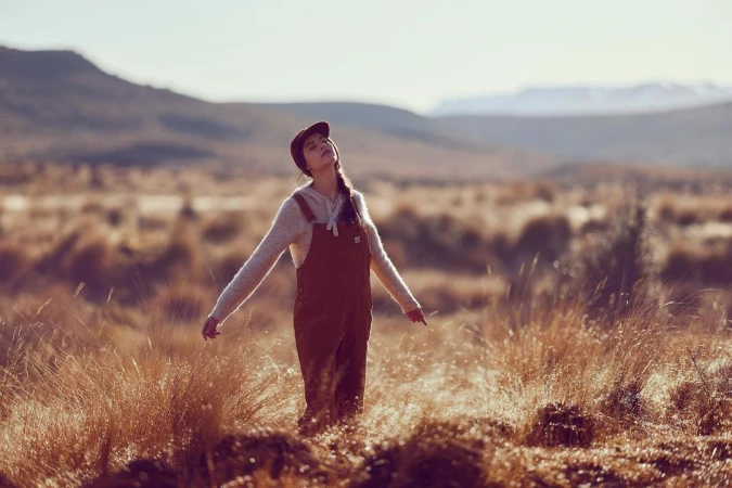 Woman in brown overalls and cap standing in a grassy field with arms outstretched under a soft hazy sky