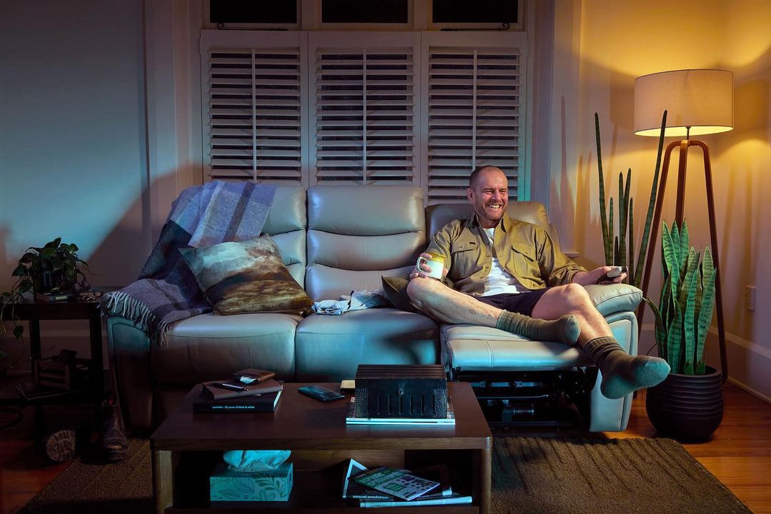 Man relaxing on a recliner sofa in a cozy living room at night, holding a mug and remote control, with soft lighting.