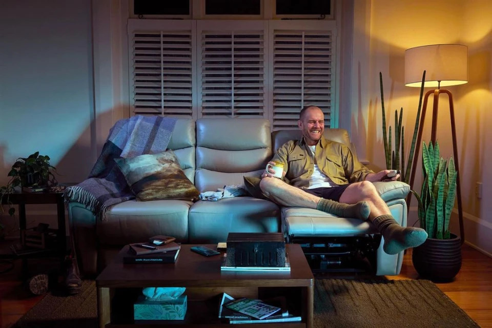 Man relaxing on a recliner sofa in a cozy living room at night, holding a mug and remote control, with soft lighting.